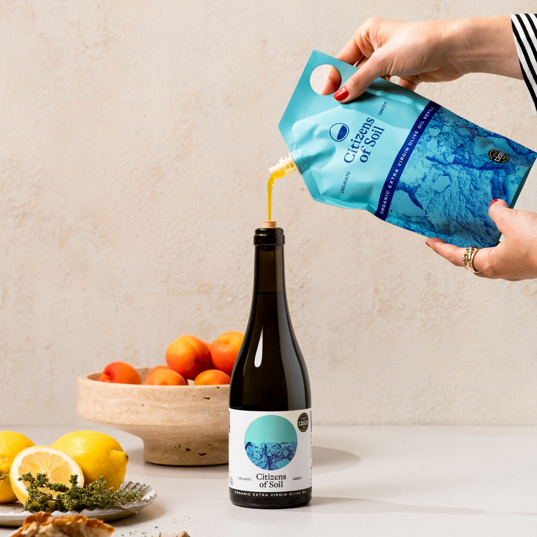 Person pouring olive oil from a 'Citizens of Soil' pouch into a bottle with lemons and oranges in the background.