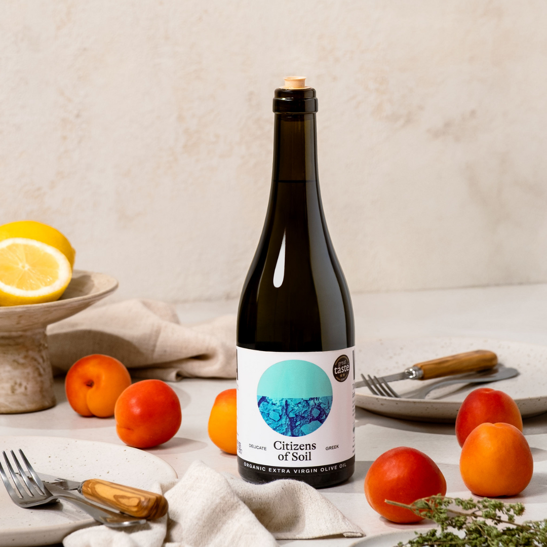 Bottle of olive oil labeled 'Citizens of Soil' on a table with fruits and cutlery.