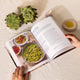 An open cookbook on a white textured fabric surface, showing a recipe page for pasta. Two hands with red nail polish hold the book open.