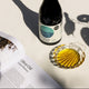 A dark glass bottle of olive oil with a cork stopper and a blue-and-white label stands on a light surface, alongside a small clear glass dish filled with golden olive oil. Strong leaf shadows are cast across the scene.