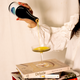 Person pouring a olive oil from a bottle into a glass on top of books with 'Drizzle' cookbook visible.